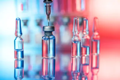 A syringe with a needle drawing the vaccine out of a vial with ampules in the background