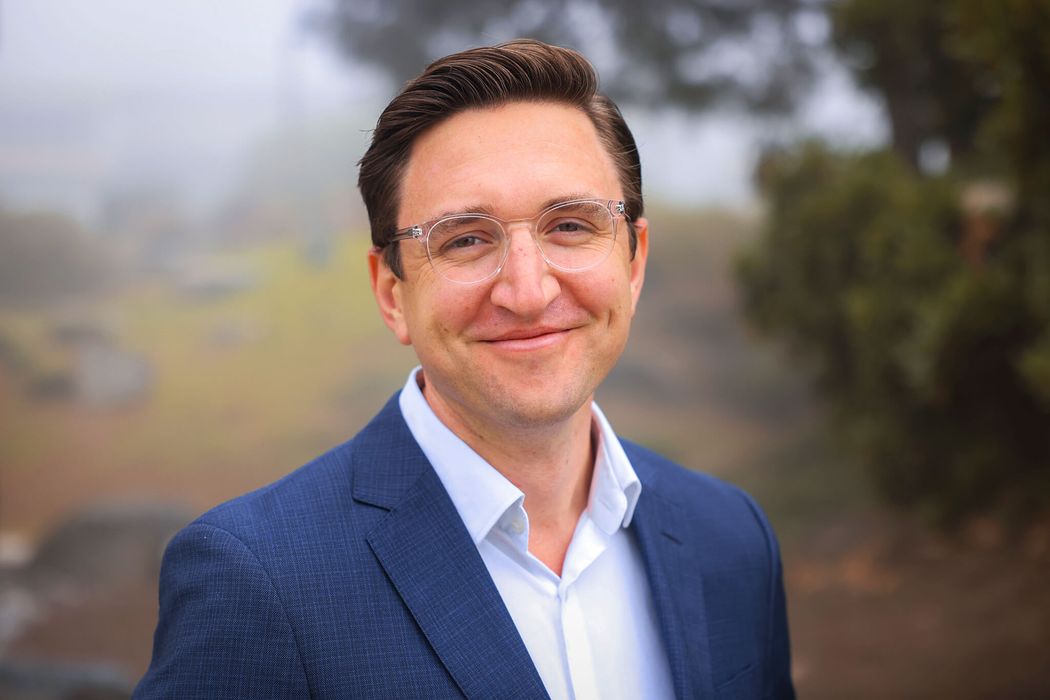Michael Bollong wears a navy blue suit jacket and glasses while standing outside. Michael Bollong wears a navy blue suit jacket and glasses while standing outside.