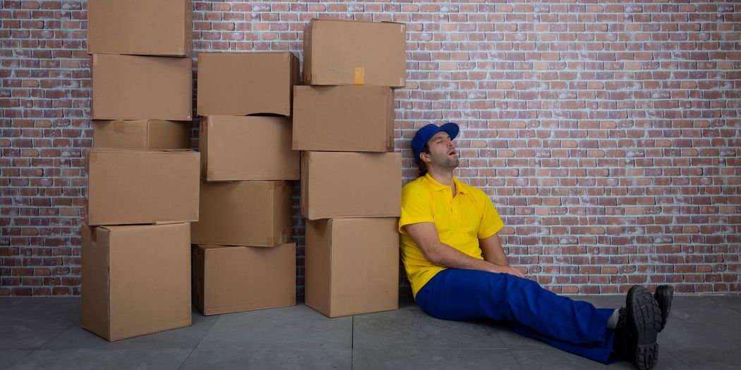 A man in yellow T-shirt, blue pants, and a cap sleeps sitting on the floor leaning against a wall, next to a pile of boxes. A man in yellow T-shirt, blue pants, and a cap sleeps sitting on the floor leaning against a wall, next to a pile of boxes.
