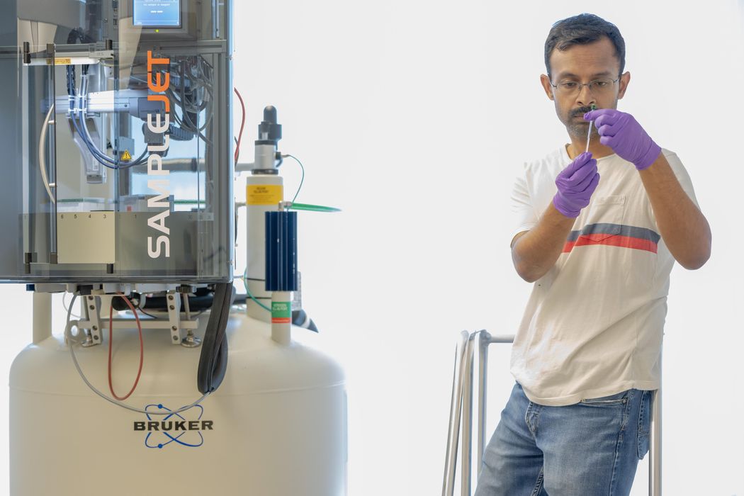 Arjun Sengupta wears purple gloves and holds an instrument. He is standing next to a nuclear magnetic resonance machine. Arjun Sengupta wears purple gloves and holds an instrument. He is standing next to a nuclear magnetic resonance machine.