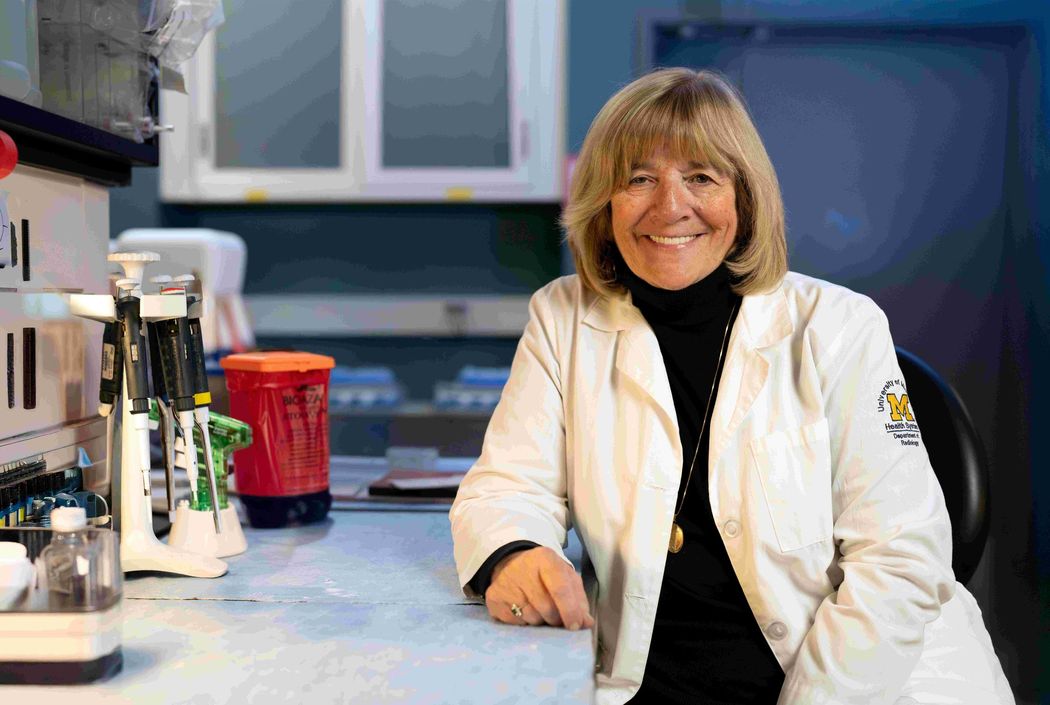 Judy Sebolt-Leopold sits at a desk wearing a white lab coat and smiles. Judy Sebolt-Leopold sits at a desk wearing a white lab coat and smiles.