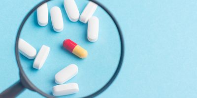 A magnifying glass highlights a red-and-yellow capsule among a scattered group of white tablets on a light blue background