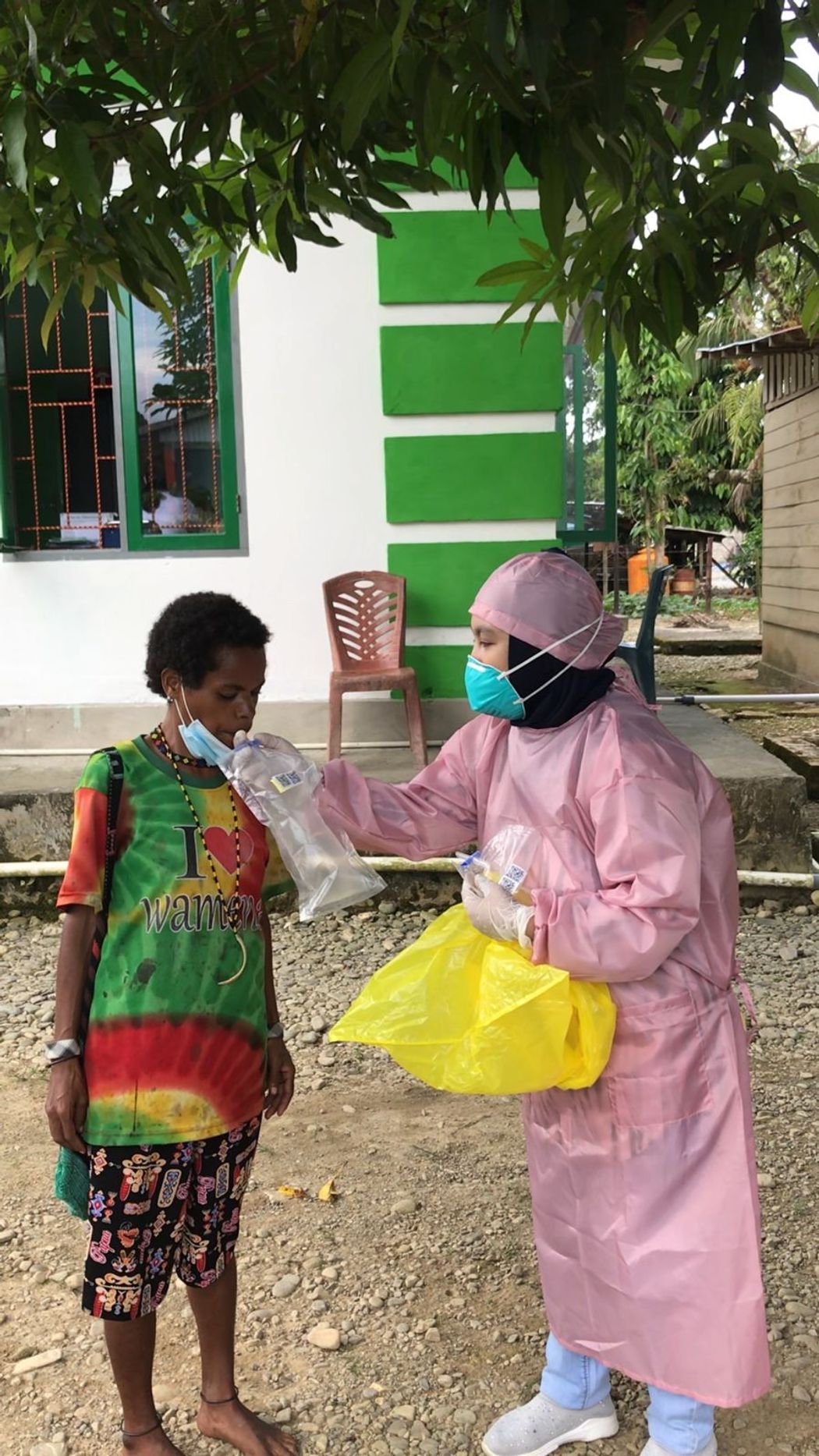 A research technician in pink personal protective equipment and mask is collecting breath samples of a person using a plastic collection bag outside a building. A research technician in pink personal protective equipment and mask is collecting breath samples of a person using a plastic collection bag outside a building.