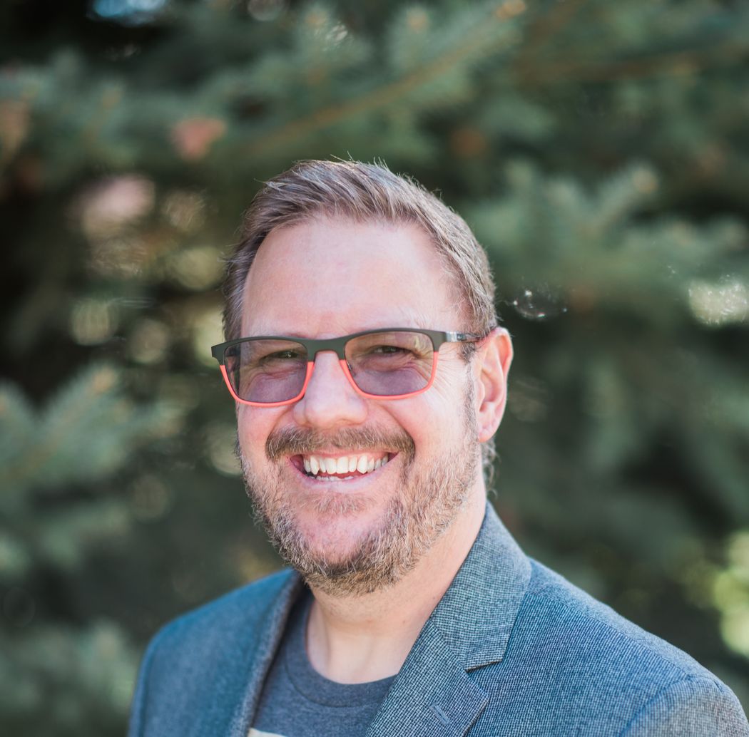 Dale Levitzke, smiling while wearing stylish rectangular glasses with red rims outdoors.
