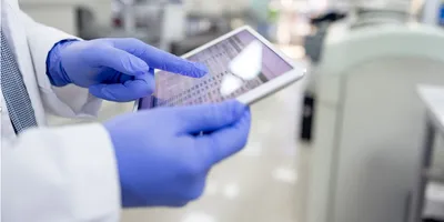 A scientist wearing blue gloves interacts with a digital tablet displaying bioanalytical data in a bright, modern laboratory setting.