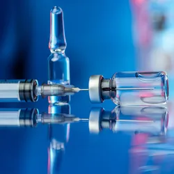 A syringe draws liquid from a glass vial, with several glass ampoules reflected on a glossy surface in the background