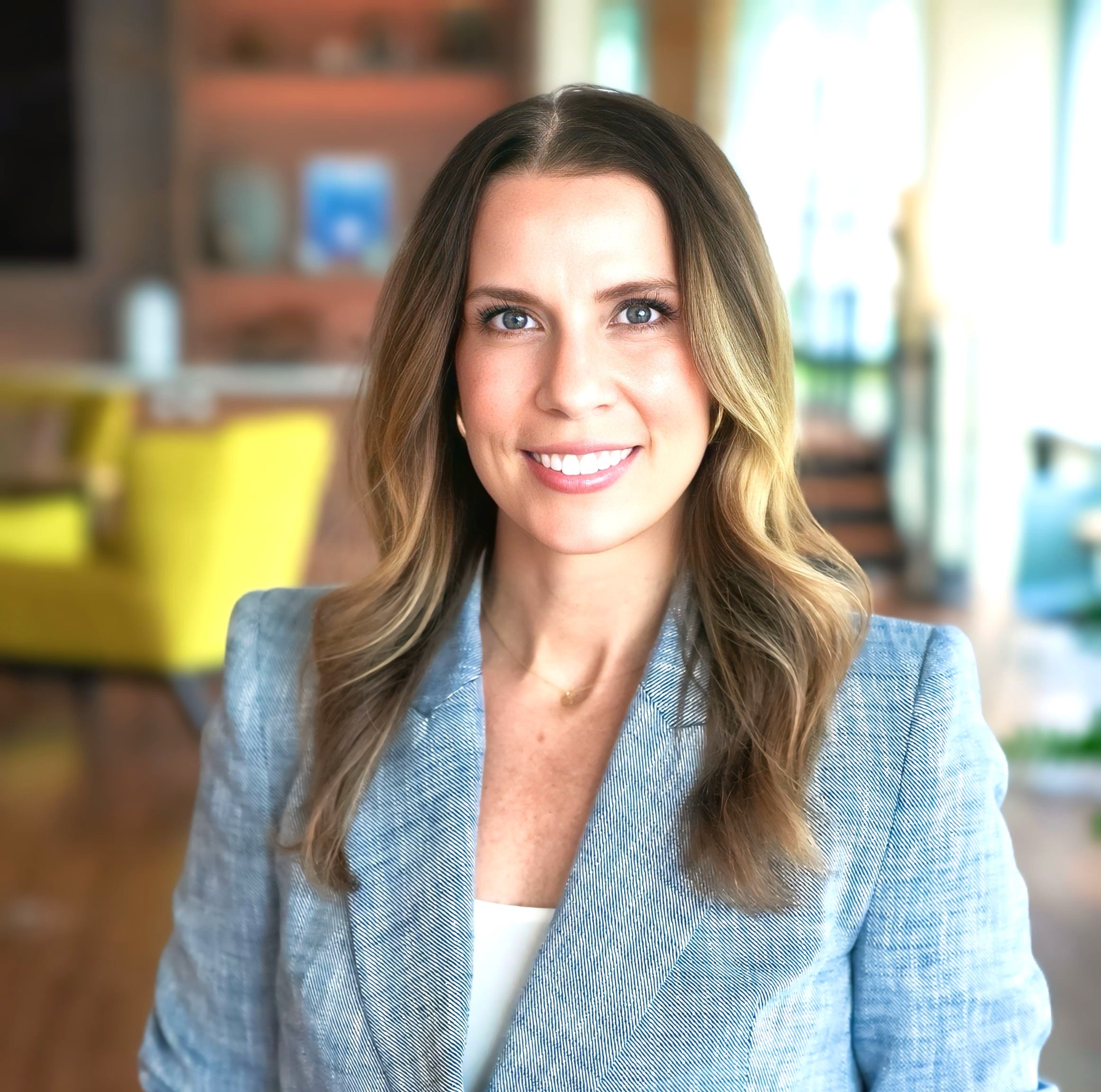 Johanna DeYoung smiling in a light blue blazer standing in a modern office setting. Johanna DeYoung smiling in a light blue blazer standing in a modern office setting.