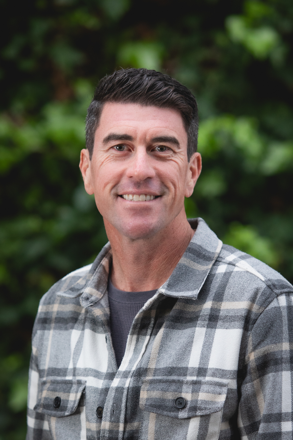 Ryan Johnson, smiling in a plaid flannel shirt while standing outdoors in front of green foliage. Ryan Johnson, smiling in a plaid flannel shirt while standing outdoors in front of green foliage.