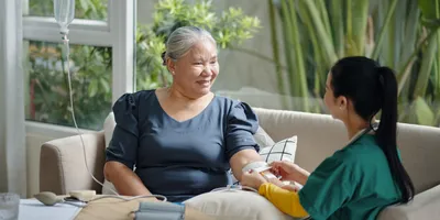 A female healthcare professional wearing green and yellow scrubs attaching an IV drip to a senior female patient at home, seated on a beige cough beside  a round wood table with plants shown in the background.  