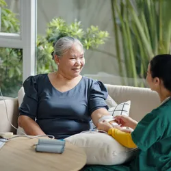 A female healthcare professional wearing green and yellow scrubs attaching an IV drip to a senior female patient at home, seated on a beige cough beside  a round wood table with plants shown in the background.  