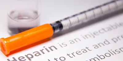 Syringe with an orange cap and glass vial containing clear liquid next to printed text explaining the medical use of heparin. 