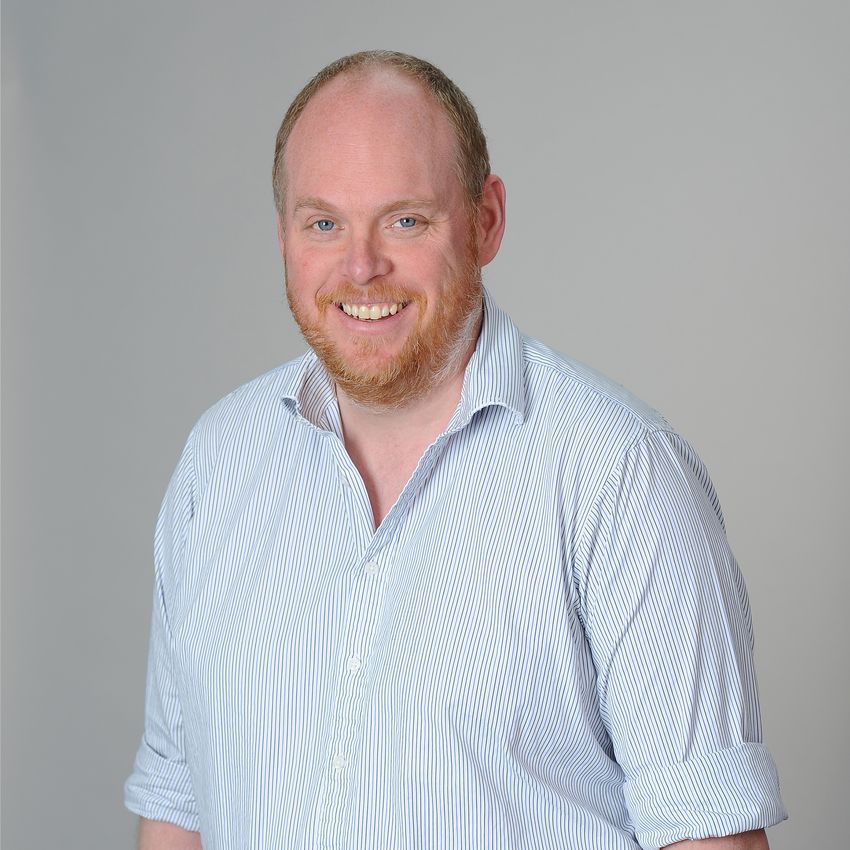 Tobias Ost, smiling in a light blue striped shirt against a plain grey background. Tobias Ost, smiling in a light blue striped shirt against a plain grey background.
