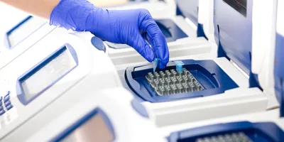 A laboratorian wearing blue gloves places a PCR sample into a real-time PCR thermocycler, the model shown is blue and white with a large digital screen.