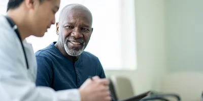 A senior gentleman wearing a blue shirt sits beside his doctor in a white lab coat as they review test results on a tablet together in a doctor’s office.