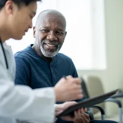 A senior gentleman wearing a blue shirt sits beside his doctor in a white lab coat as they review test results on a tablet together in a doctor’s office.