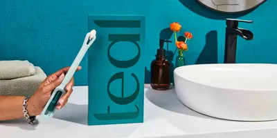 A hand holds the Teal Wand in front of its package and on the counter next to a sink.