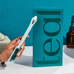 A hand holds the Teal Wand in front of its package and on the counter next to a sink.
