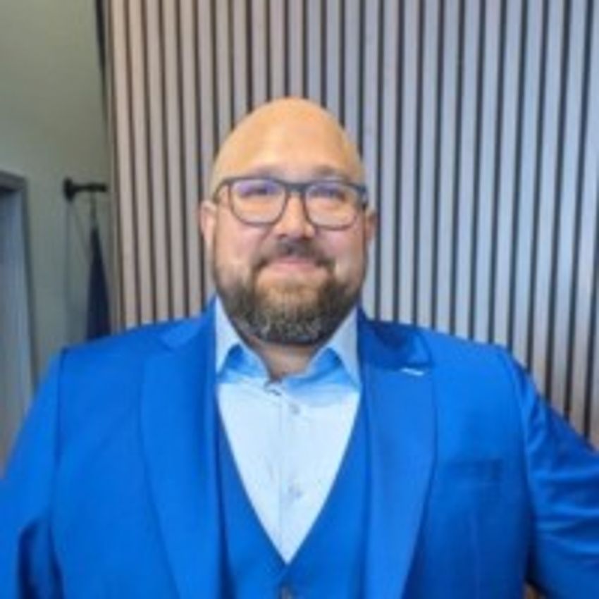 Kristofer Mussar in a bright blue suit and glasses standing in front of a modern wooden panel wall. Kristofer Mussar in a bright blue suit and glasses standing in front of a modern wooden panel wall.