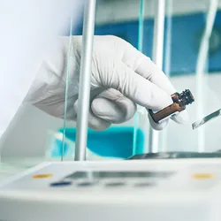 A gloved hand weighs a powdered substance using a spatula and a precision balance inside a laboratory.