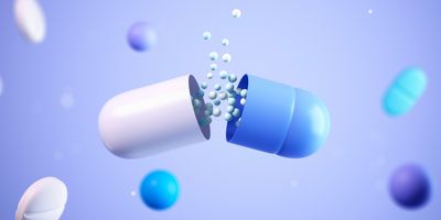 A 3D illustration of a capsule splitting open to release small white and blue granules, surrounded by floating pills and spheres on a light blue background.