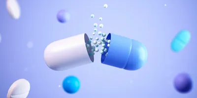 A 3D illustration of a capsule splitting open to release small white and blue granules, surrounded by floating pills and spheres on a light blue background.