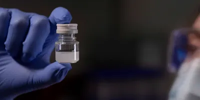 A hand wearing a blue glove holds a small clear bottle with a whitish liquid on a black background. 