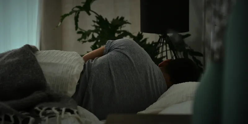 A person lying on their side in bed under a blanket, with dim lighting and indoor plants in the background, suggesting a scene of rest or fatigue.