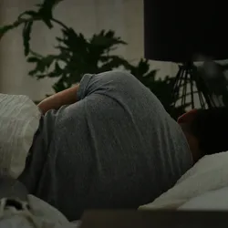 A person lying on their side in bed under a blanket, with dim lighting and indoor plants in the background, suggesting a scene of rest or fatigue.
