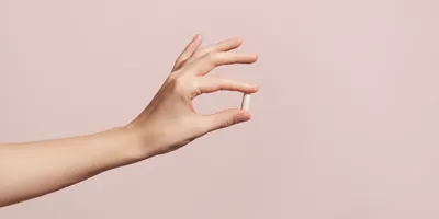 A hand holding a small white capsule between the thumb and index finger against a soft pink background.