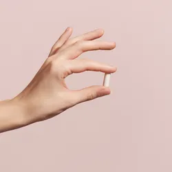 A hand holding a small white capsule between the thumb and index finger against a soft pink background.