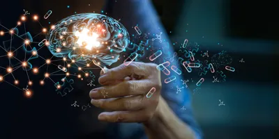 A person’s hand holds up a 3D rendering of a brain with a glowing puzzle piece in the middle and a network of lines and dots, molecules, and drug capsules surrounding it. 