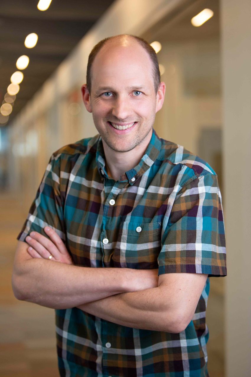 Ryan Larson smiles wearing a blue, white, and brown checkered shirt with his arms crossed.