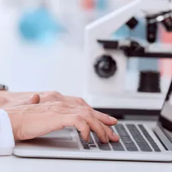 Close-up of a scientist’s hands typing on a laptop next to a microscope in a laboratory setting.