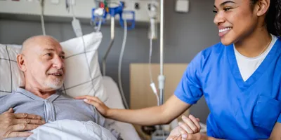 A senior man lies in a hospital bed, smiling and interacting with a clinical trial worker.   