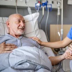 A senior man lies in a hospital bed, smiling and interacting with a clinical trial worker.   