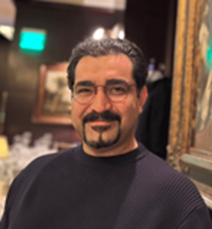Arman Zaribafiyan, wearing a dark sweater and smiling at the camera in an indoor setting with warm lighting and framed artwork in the background.