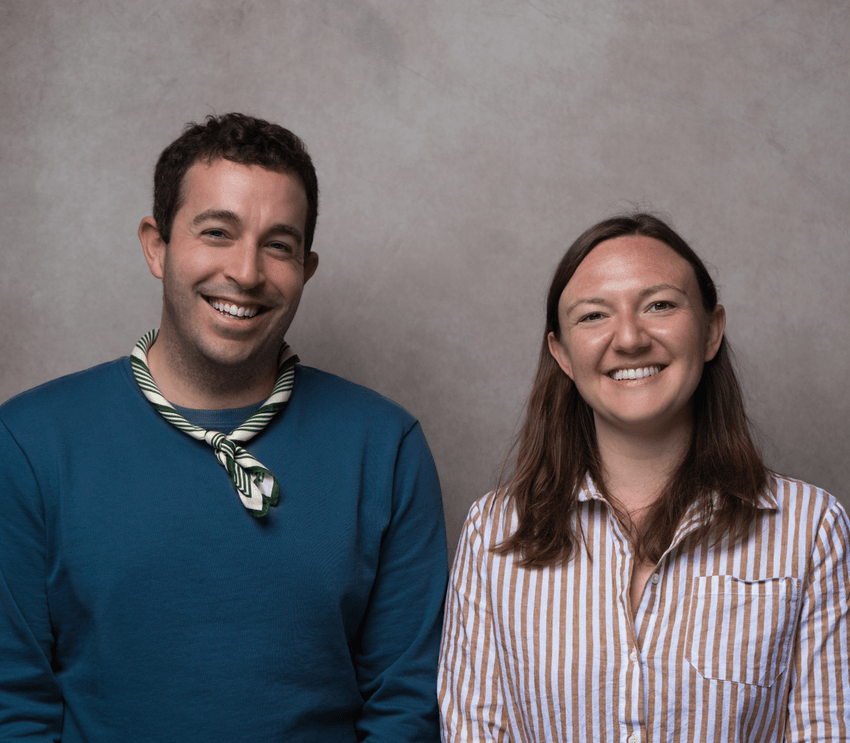 Robert DiFazio in a blue shirt with a striped scarf and Juliana Hilliard in a striped button-down, smiling together in front of a gray backdrop.