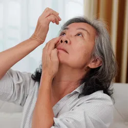 An older woman using eye drops while sitting on a couch.