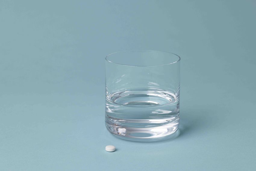A photo of a clear glass next to a white pill on a blue background.