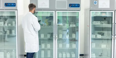 A laboratory technician in a white coat inspects temperature-controlled storage refrigerators filled with labeled scientific supplies.