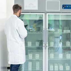 A laboratory technician in a white coat inspects temperature-controlled storage refrigerators filled with labeled scientific supplies. 