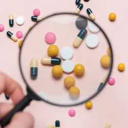 A hand holding a magnifying glass focuses on assorted colorful pills and capsules scattered on a light background, symbolizing the close examination and detection of pharmaceutical impurities.