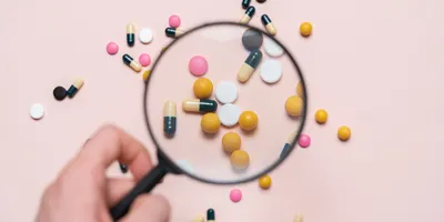 A hand holding a magnifying glass focuses on assorted colorful pills and capsules scattered on a light background, symbolizing the close examination and detection of pharmaceutical impurities.