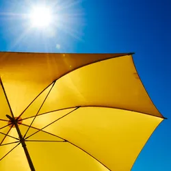 A bright yellow umbrella is shown under the sun and a blue sky.