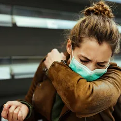 Sick woman wearing a face mask and coughing into elbow during COVID-19 pandemic.