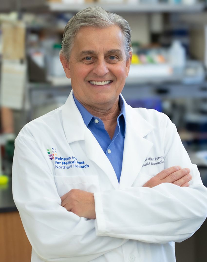 Image of Kevin Tracey smiling with his arms crossed, wearing a white lab coat over a blue dress shirt