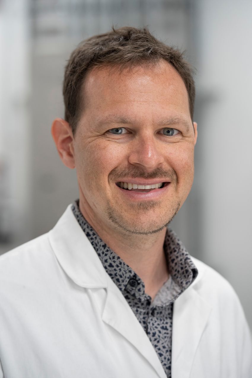 Headshot of Paul Seidler smiling, wearing a white lab coat over a black and gray patterned shirt