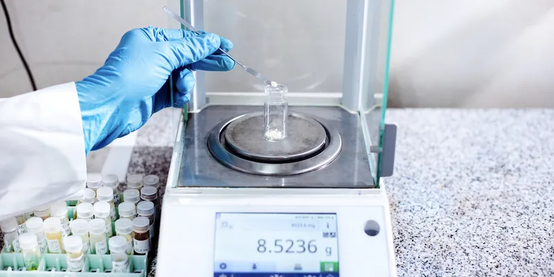 Scientist weighing a laboratory sample using a four-decimal analytical balance in a quality control setting.
