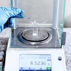 Scientist weighing a laboratory sample using a four-decimal analytical balance in a quality control setting.