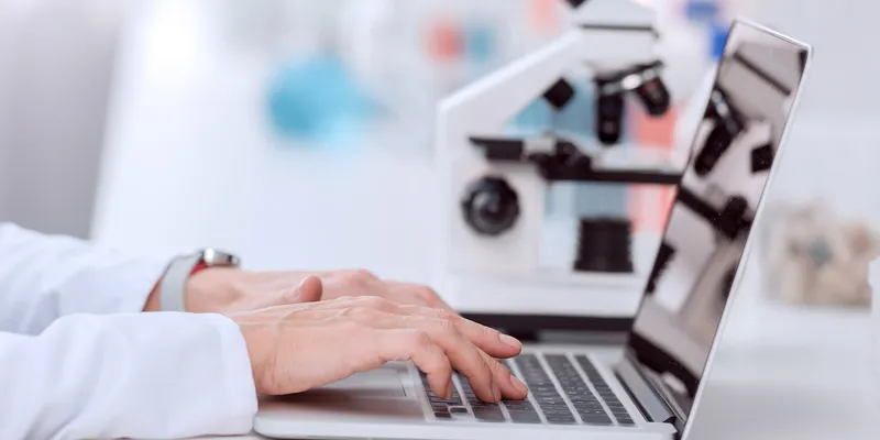 Close-up of a scientist’s hands typing on a laptop next to a microscope in a laboratory setting.